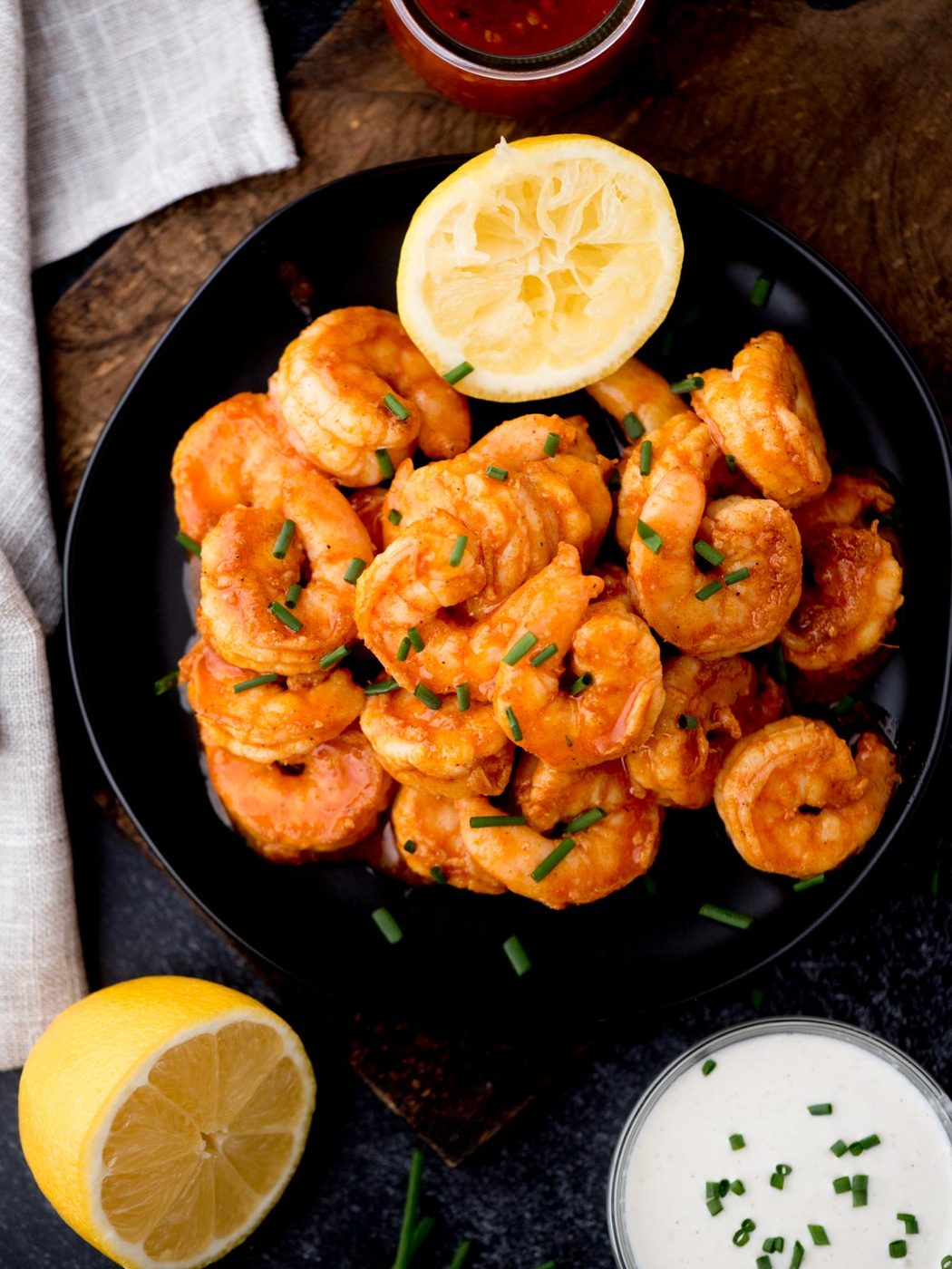 A black plate filled with cooked shrimp garnished with chopped chives. A halved lemon is placed on top of the shrimp. There is a small bowl of creamy dressing and a glass of red sauce nearby. A whole lemon and light cloth are on the side.