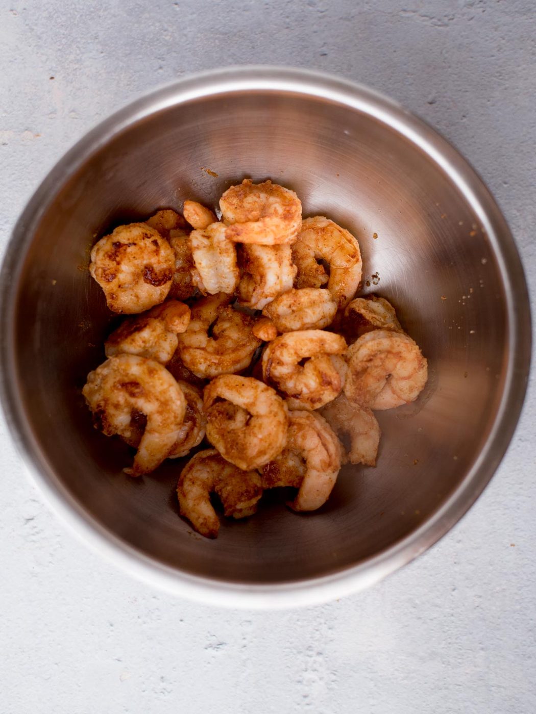 A stainless steel bowl filled with cooked, golden-brown shrimp on a light gray background. The shrimp appear seasoned and slightly crispy.