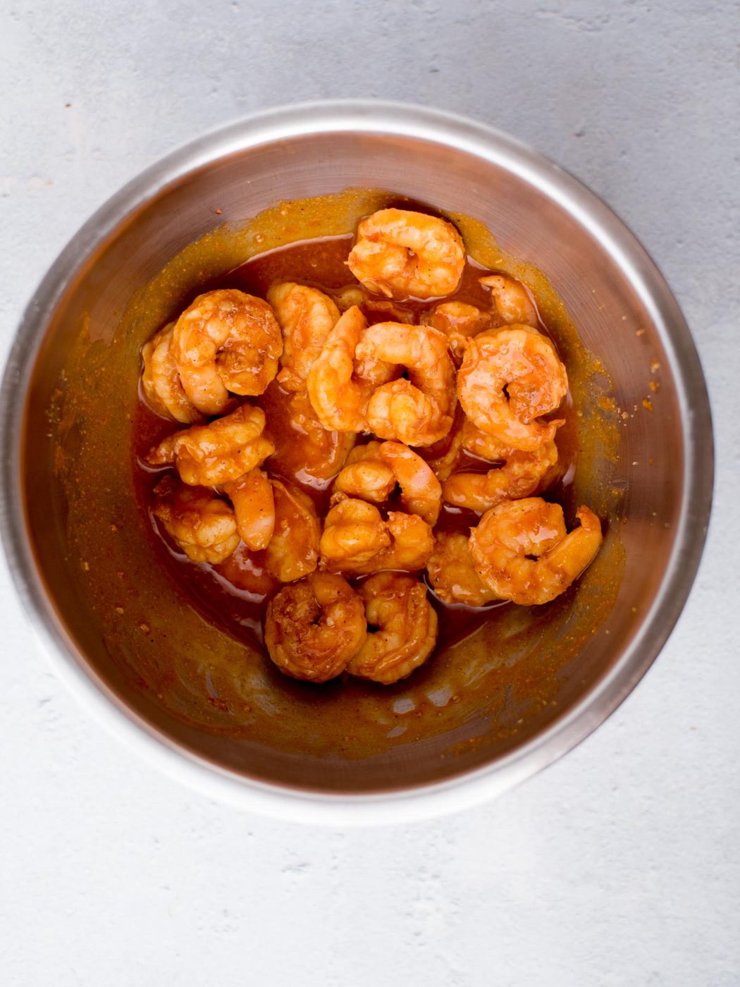 A stainless steel bowl filled with shrimp, coated in a rich, tangy sauce. The shrimp are plump and glistening, arranged in a spiral pattern against a light gray background.