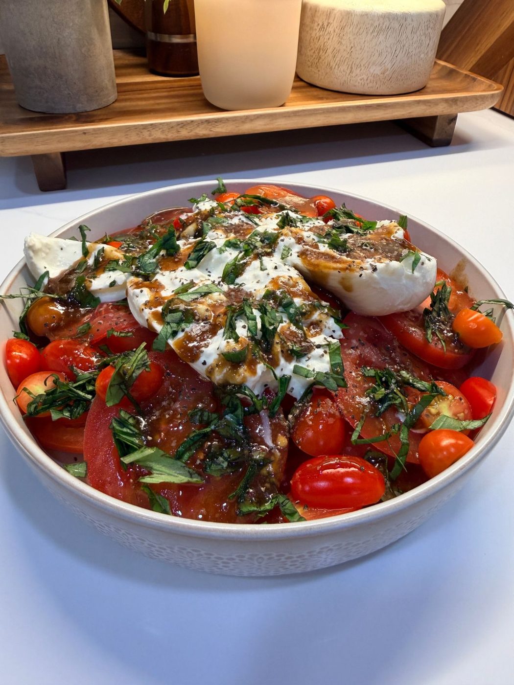 A bowl of caprese salad with sliced tomatoes, cherry tomatoes, fresh mozzarella, chopped basil, and a drizzle of balsamic dressing, set on a white countertop.