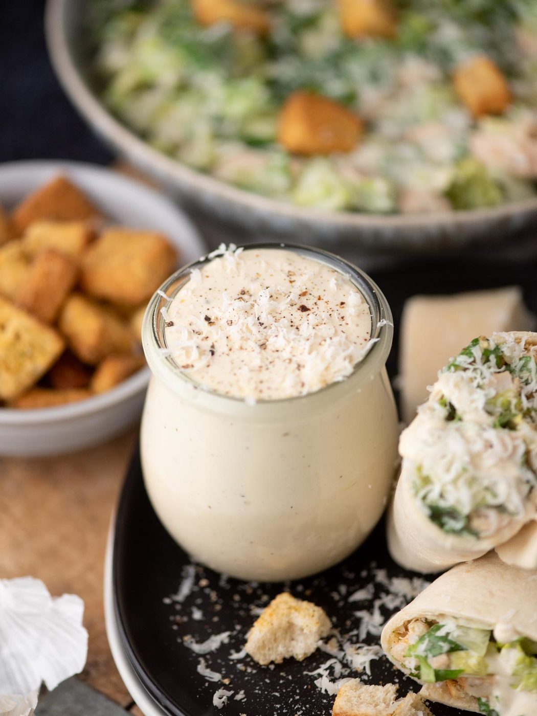 A jar of creamy Caesar salad dressing is surrounded by croutons, a wrap filled with salad, and a bowl of Caesar salad in the background. Grated cheese is sprinkled on top of the dressing and the wrap.