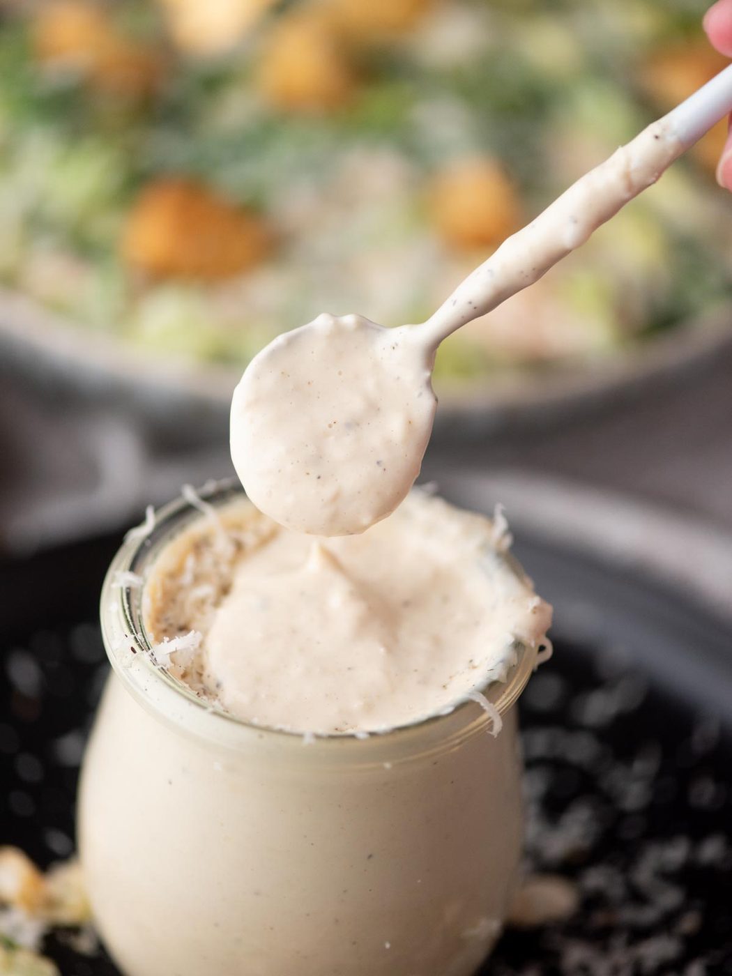 A close-up of a jar filled with creamy Caesar salad dressing, with a spoon lifting some of the dressing above the jar. The background shows a blurred bowl of salad.