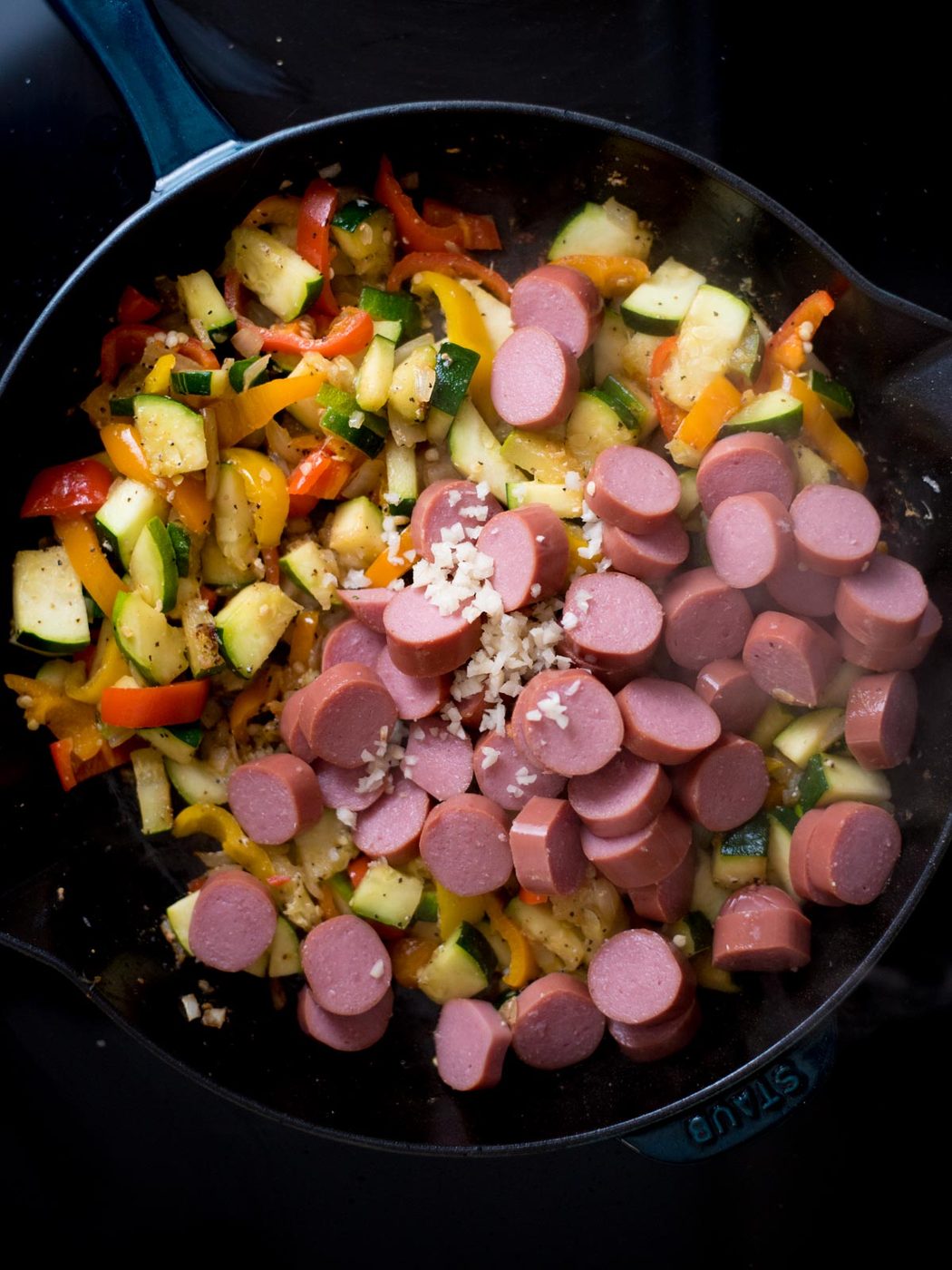 Raw cut up veggie and sausage pieces with a splash of broth inside a cast iron skillet.
