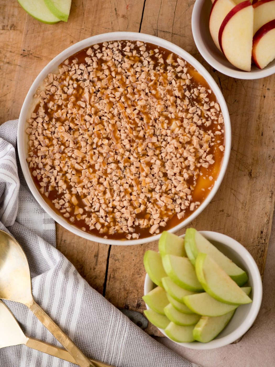 caramel apple dip in a white dish on a wooden board surrounded by bowls of sliced apples, a dish towel, and gold utensils