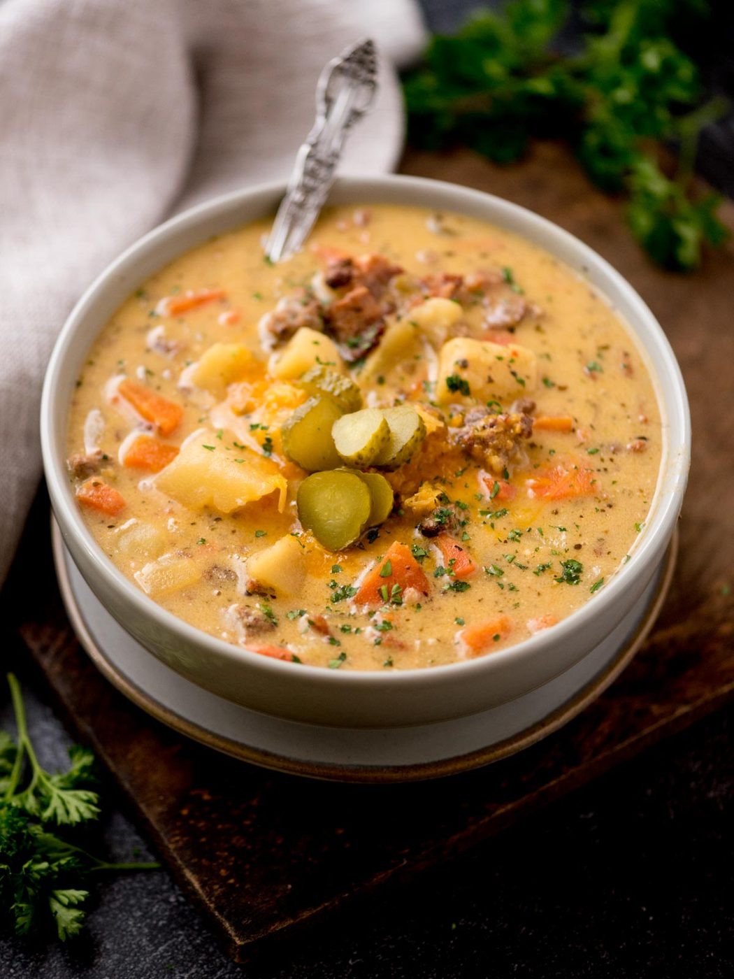 cheeseburger soup in a grey bowl with a spoon topped with chopped pickles and cheese. It is on a brown board surrounded by a kitchen towel and fresh parsley.
