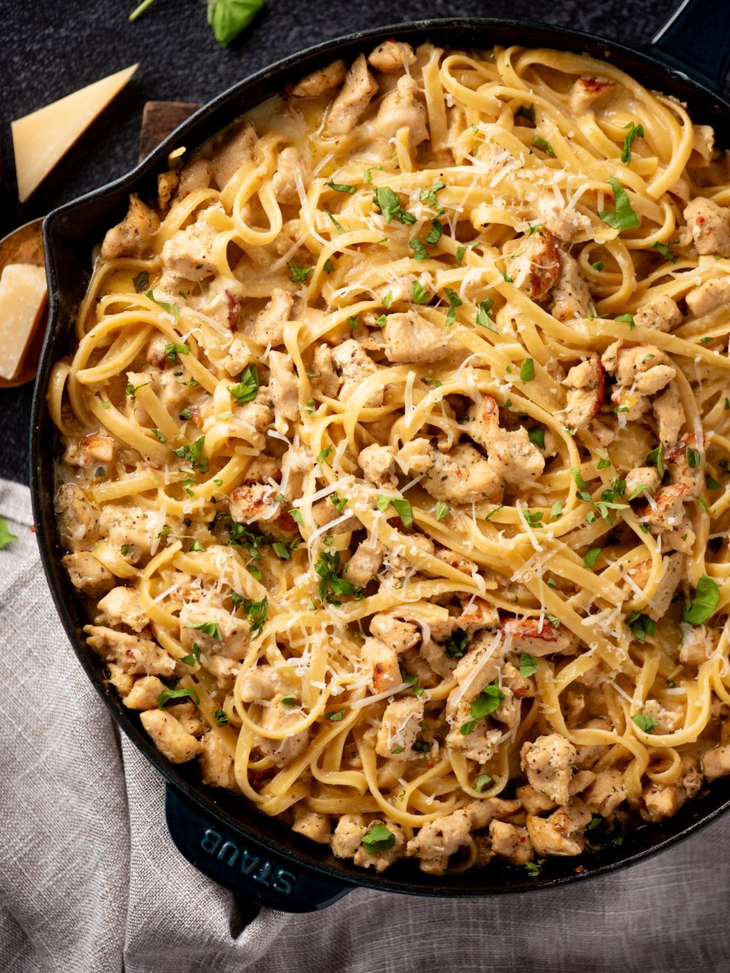 A skillet filled with creamy pasta topped with pieces of cooked chicken, garnished with grated cheese and chopped fresh herbs, sits on a gray cloth with blocks of parmesan nearby.