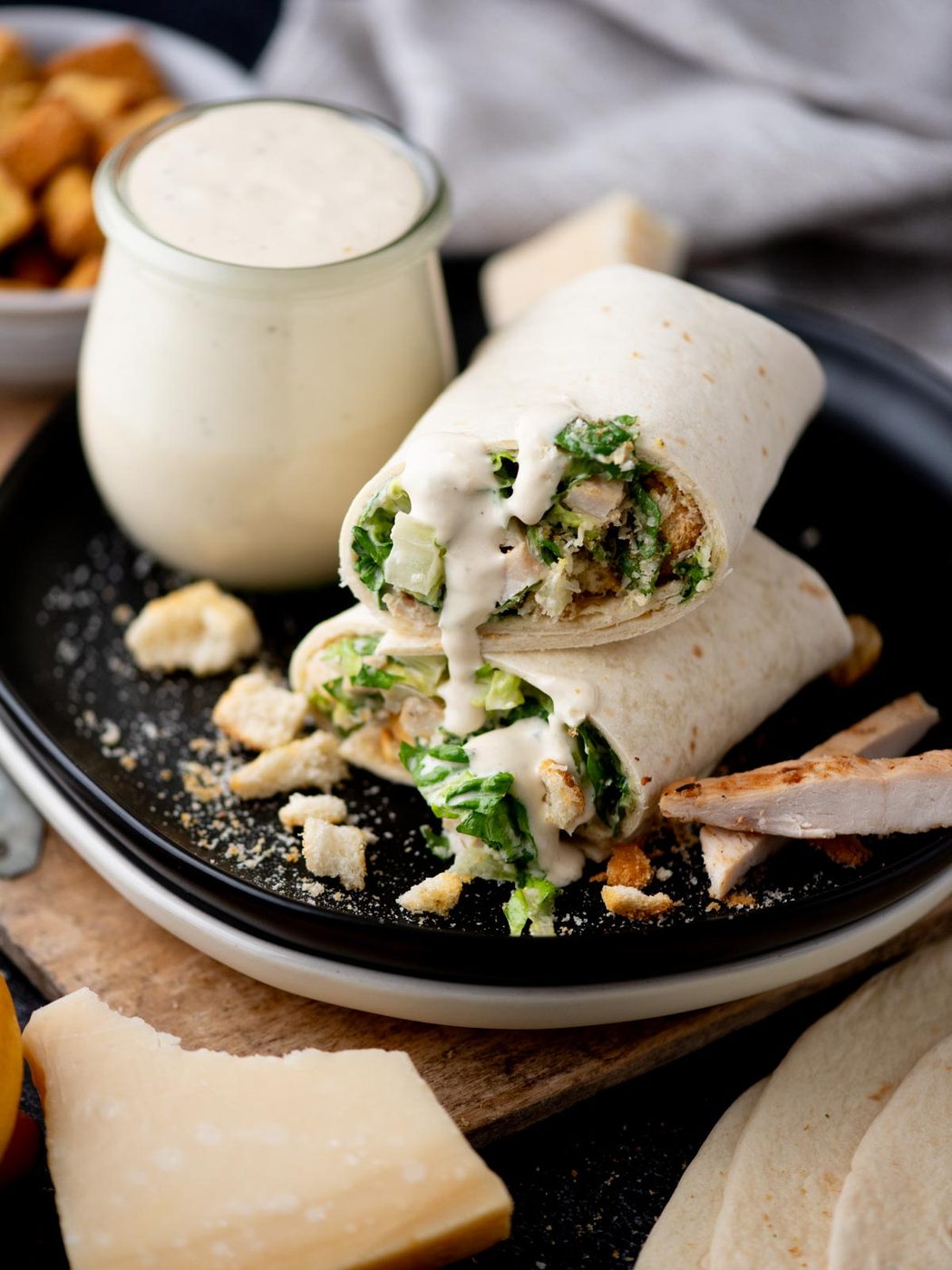 A Caesar salad wrap cut in half sits on a black plate, filled with lettuce, chicken, croutons, and creamy Caesar dressing, with extra dressing in a jar nearby and sprinkled cheese on the plate.