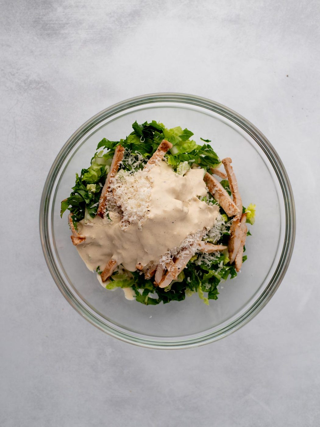 A glass bowl filled with chopped romaine lettuce, grilled chicken strips, grated cheese, and a generous amount of creamy dressing on top, set against a light gray background.