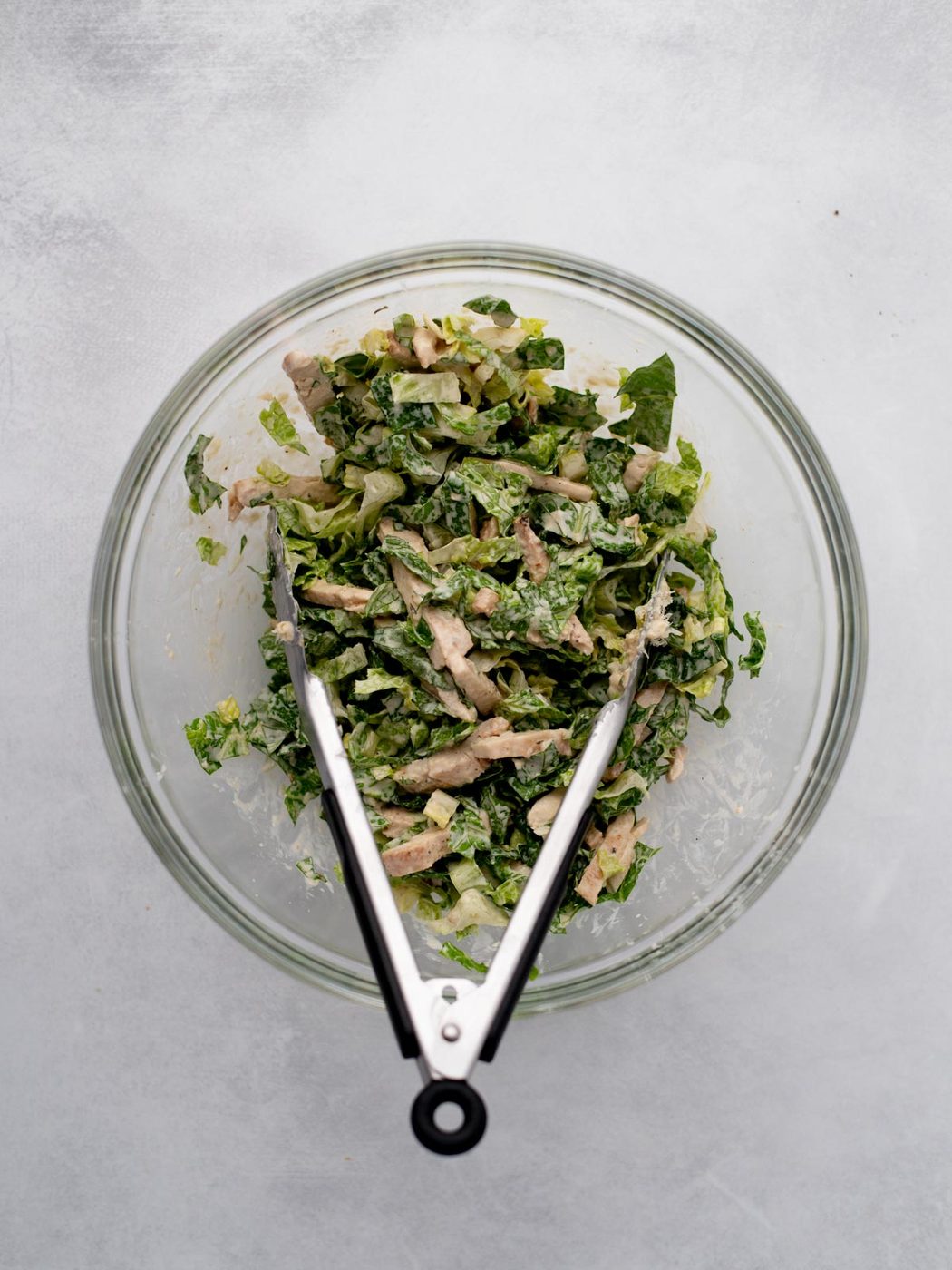 A glass bowl filled with chopped salad greens and pieces of cooked chicken, with metal tongs resting inside the bowl, on a light gray surface.