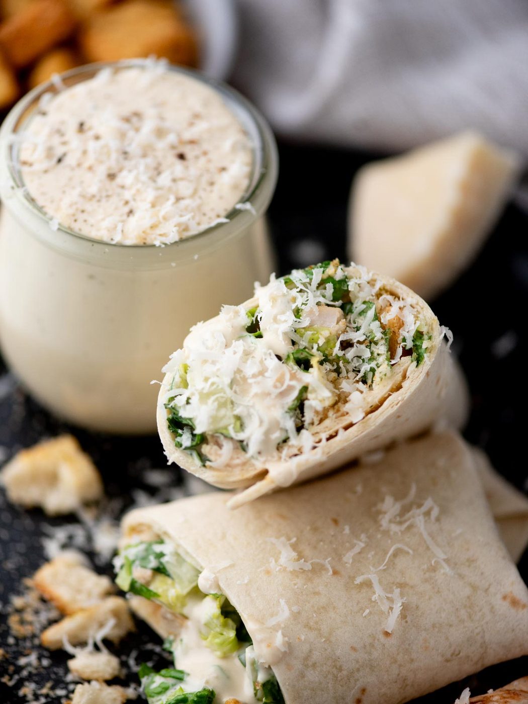 A close-up of a Caesar salad wrap cut in half, filled with lettuce, chicken, and grated cheese, with a jar of creamy Caesar dressing and croutons in the background.
