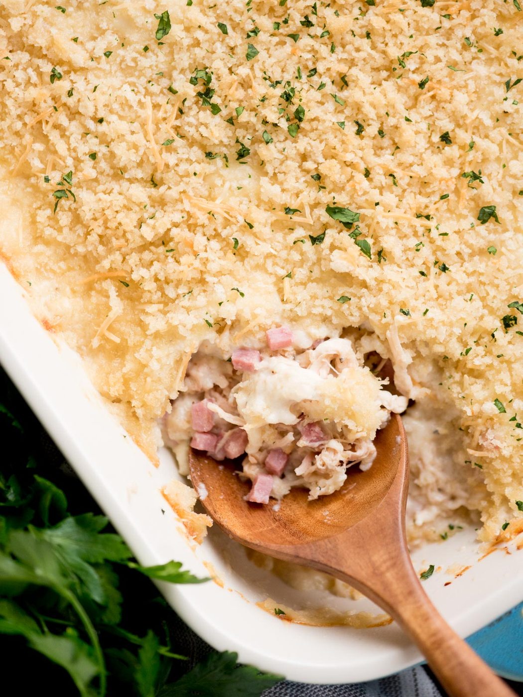 chicken cordon bleu casserole in a baking dish with a serving spoon inside. It is surrounded by fresh parsley and a kitchen towel.