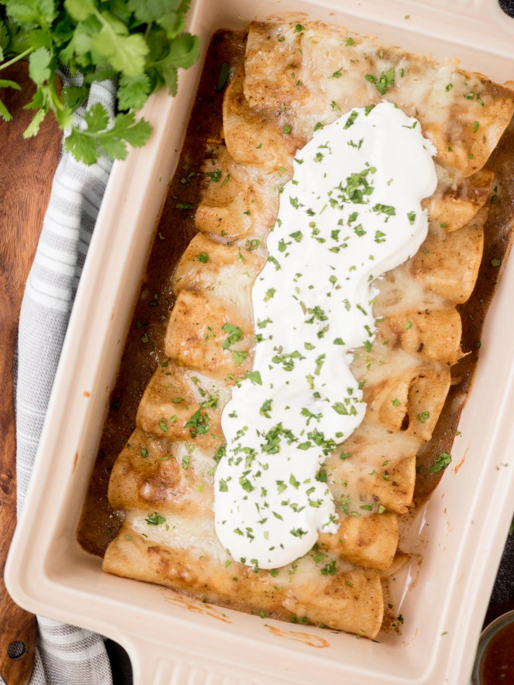 chicken enchiladas topped with sour cream and cilantro in a baking dish surrounded by serving utensils and fresh cilantro