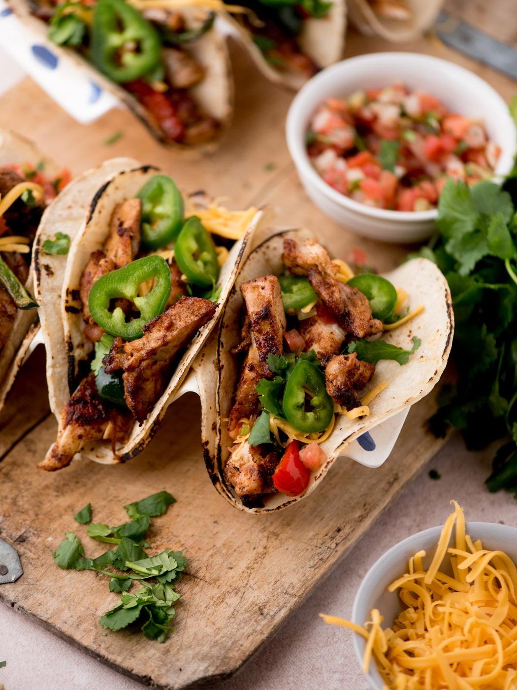 Three grilled chicken tacos, inspired by Texas chicken fajitas, topped with sliced jalapeños, shredded cheese, and diced tomatoes on a wooden board, surrounded by fresh cilantro, extra cheese, and a bowl of pico de gallo.