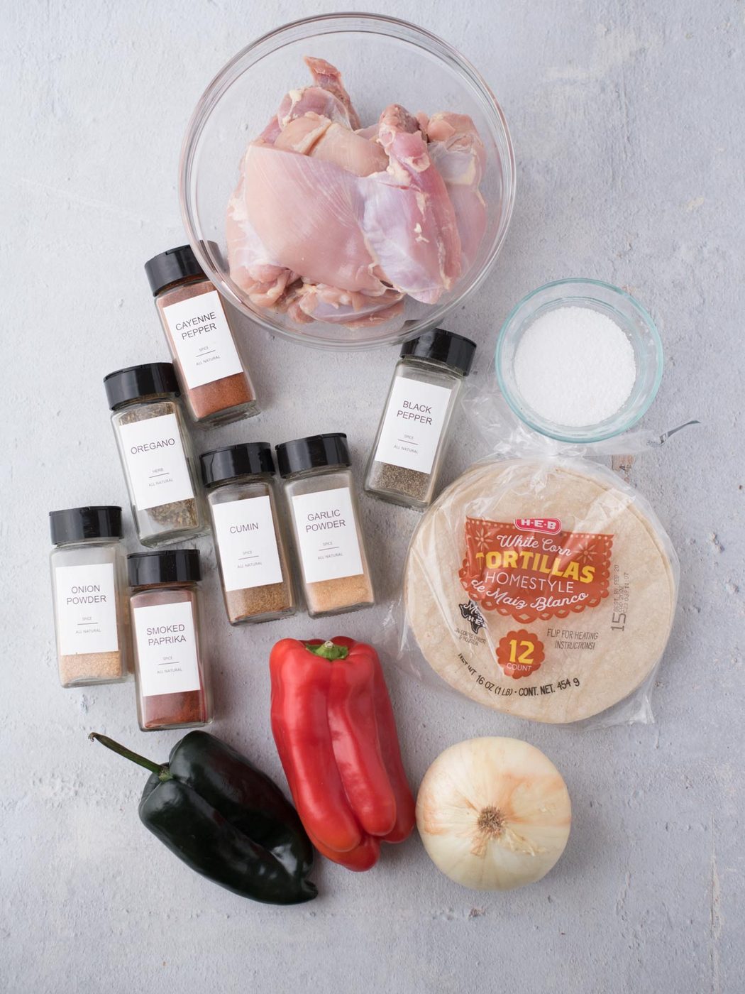 Overhead view of raw chicken in a bowl, a stack of tortillas, salt, red and poblano peppers, onion, and spice jars—well-balanced ingredients for straightforward chicken fajitas or classic Texas chicken fajitas.