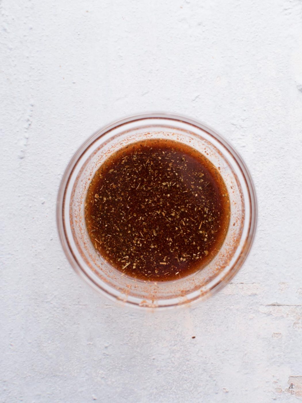 A glass bowl containing a brown, herb-speckled liquid sits on a light, textured surface. This flavorful mixture appears to be a marinade or dressing, suited to making straightforward chicken fajitas.
