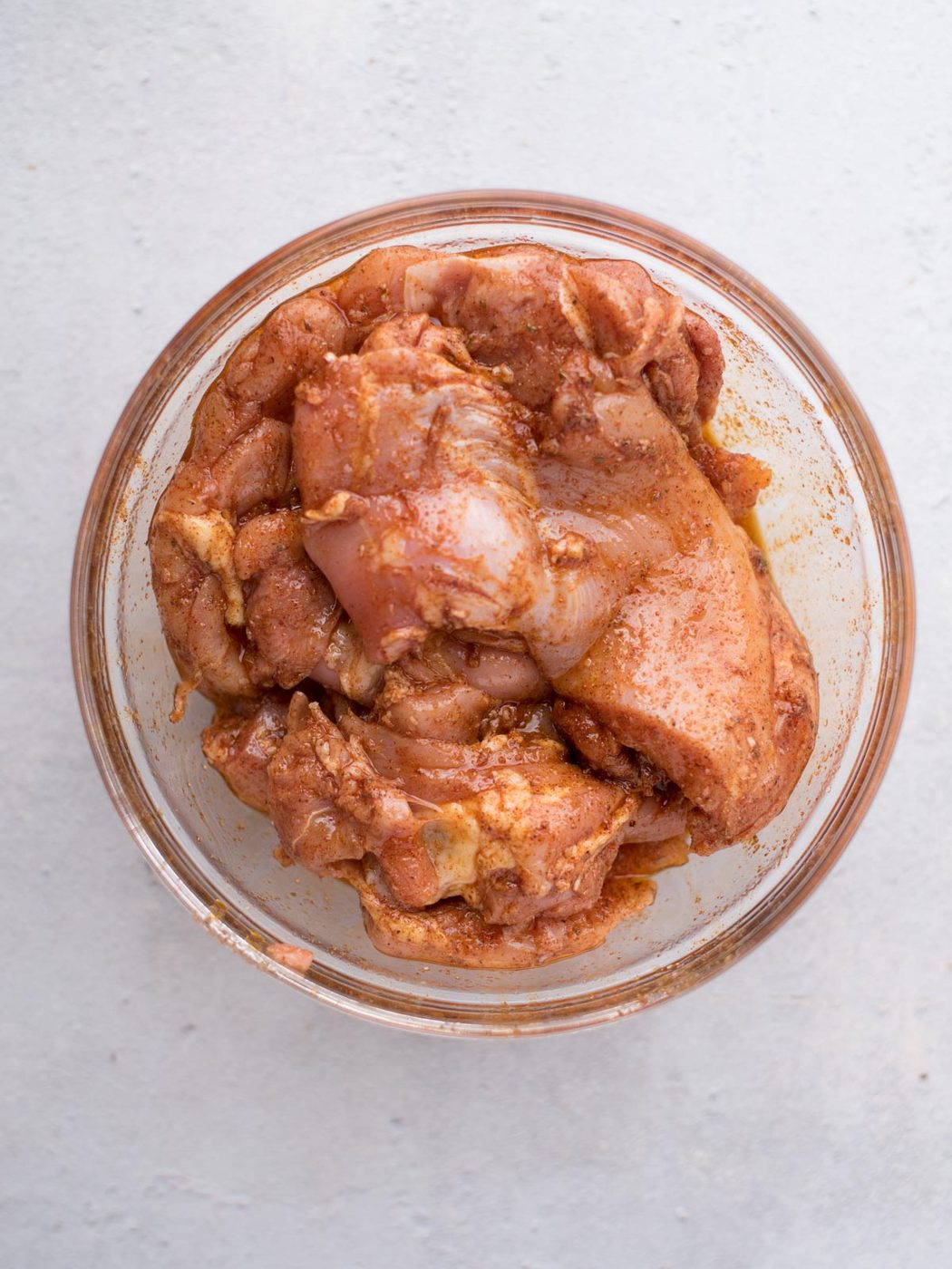 A glass bowl filled with raw chicken pieces coated in a reddish-brown marinade, suited to making straightforward chicken fajitas, sits on a light-colored surface.
