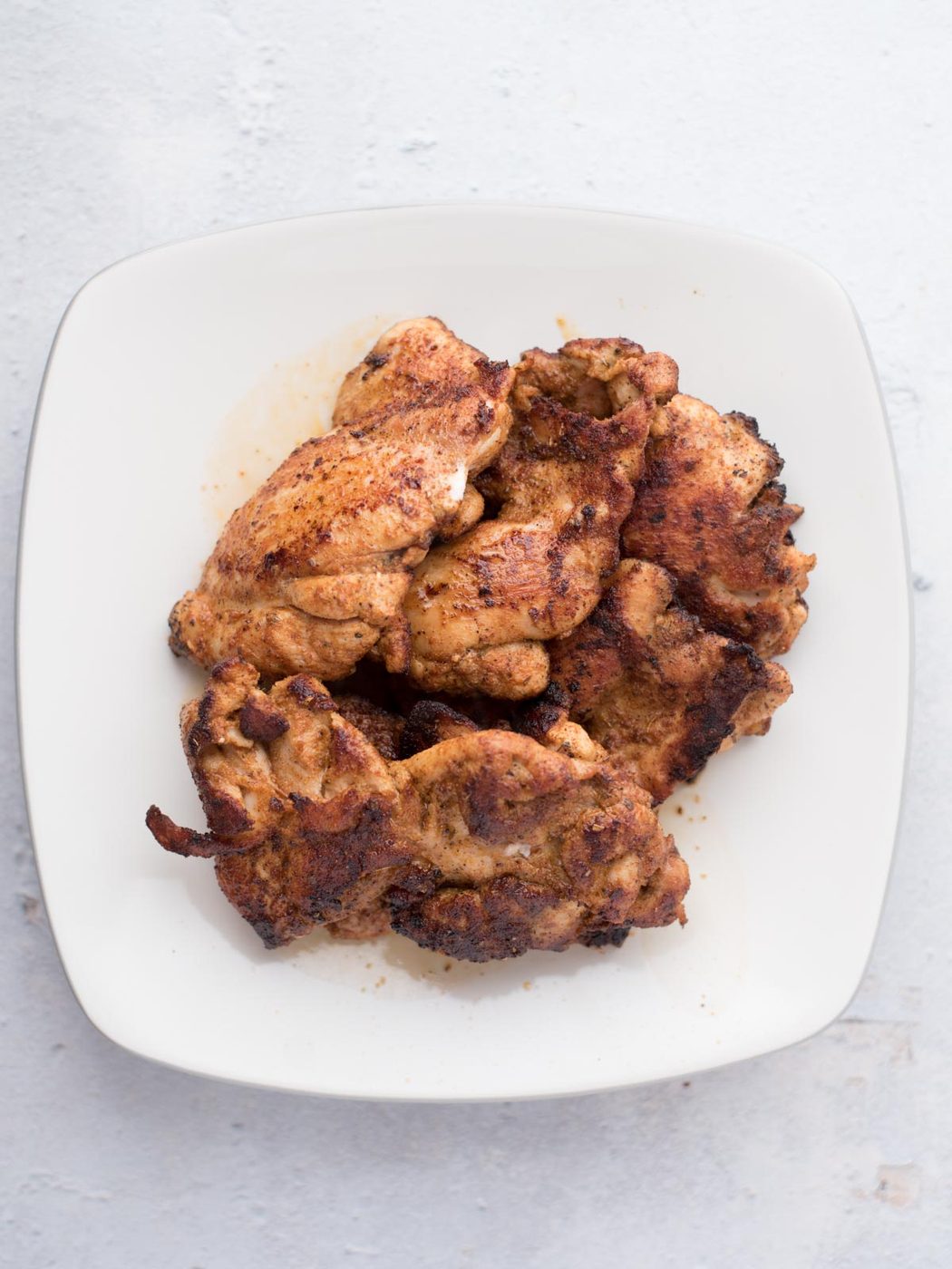 A white plate with several pieces of cooked, seasoned chicken thighs stacked on it, suited to making straightforward chicken fajitas, is placed on a light-colored surface.