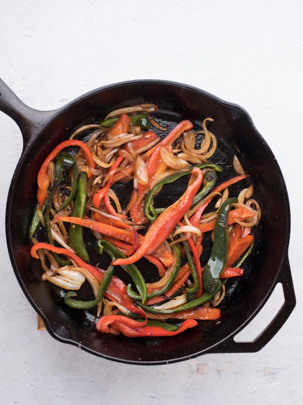 Sautéed sliced red and green bell peppers with onions in a black cast iron skillet, the well-balanced filling for straightforward chicken fajitas, viewed from above on a white background.