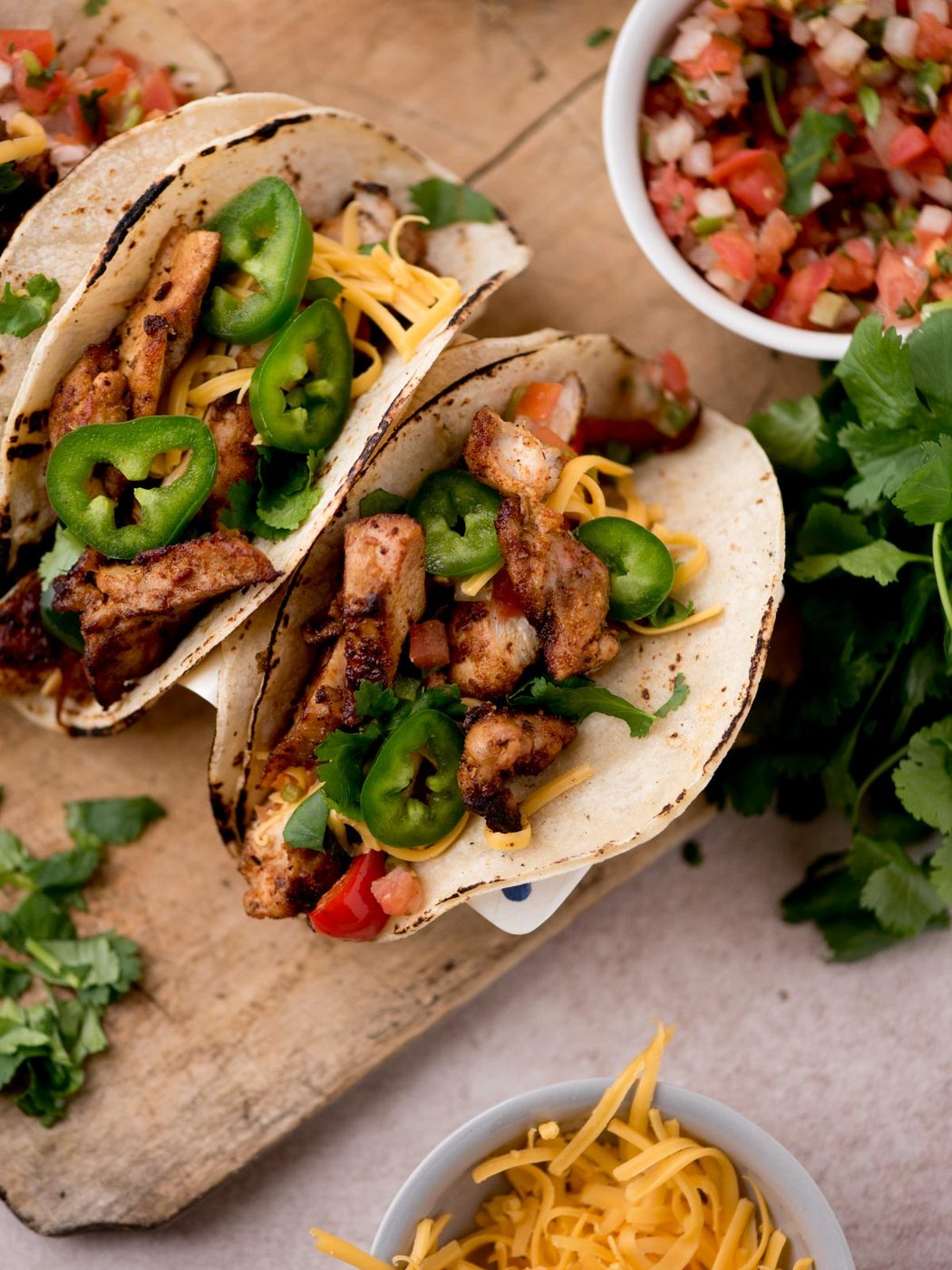 Three grilled chicken tacos inspired by Texas chicken fajitas, topped with sliced jalapeños, shredded cheese, and diced tomatoes on corn tortillas. Served on a wooden board with fresh cilantro, pico de gallo, and extra cheese.