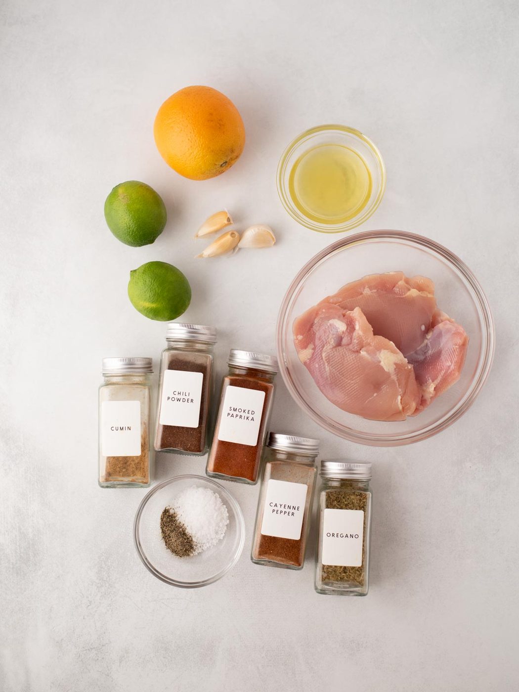 Raw chicken breast in a glass bowl, surrounded by spices in jars (cumin, chili powder, smoked paprika, cayenne pepper, oregano), black pepper, salt, garlic, lime, orange, and a small bowl of oil on a white surface.