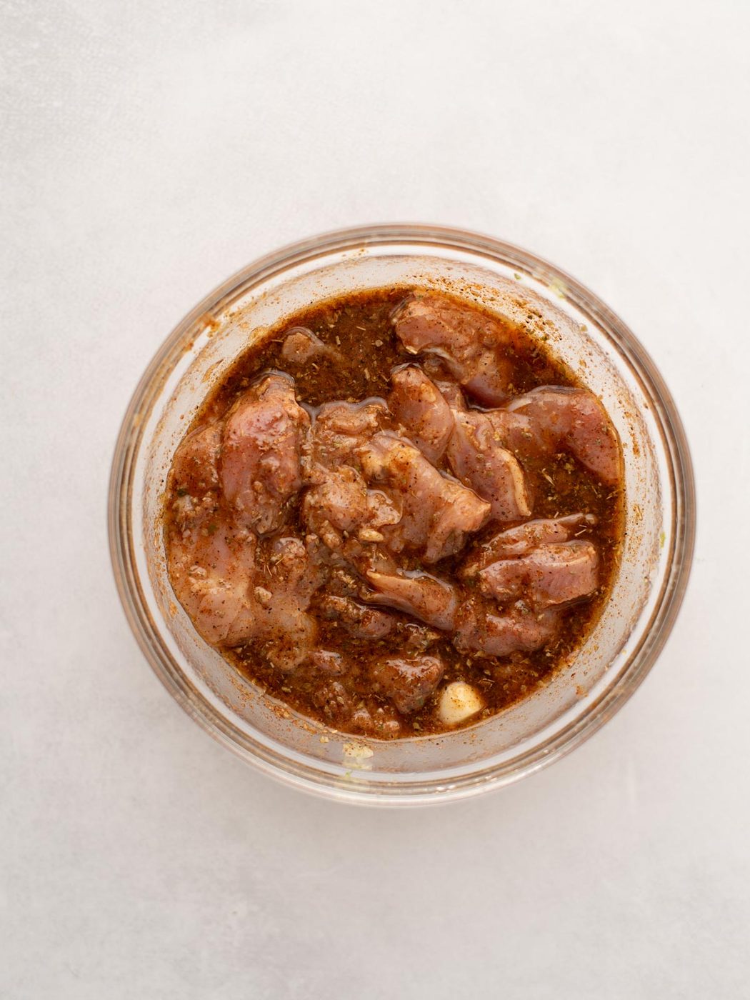 A glass bowl filled with pieces of raw chicken marinating in a dark, seasoned sauce, placed on a light-colored surface.