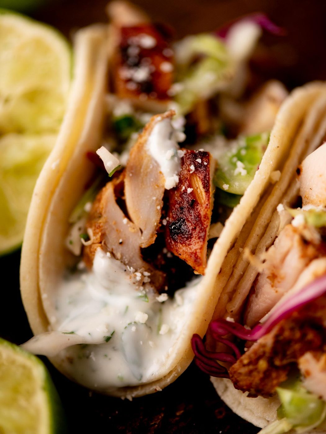 Close-up of a chicken taco in a soft tortilla, topped with creamy white sauce, shredded lettuce, and grilled chicken pieces. Lime wedges are visible in the background.