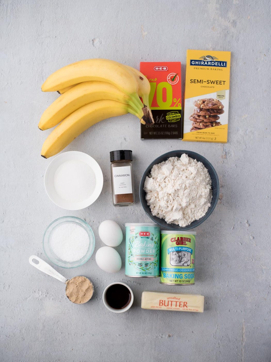 Overhead view of ingredients for baking: three bananas, two chocolate bars, bowl of flour, bowl of sugar, cinnamon, salt, two eggs, baking powder, baking soda, brown sugar, vanilla extract, and a stick of butter.