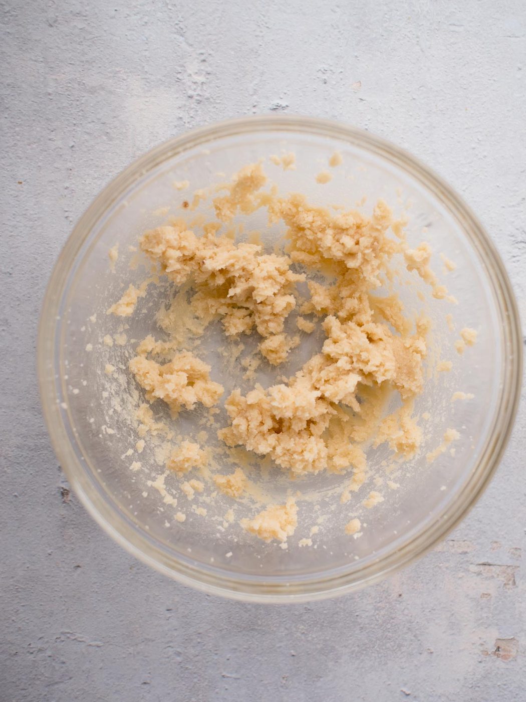 A glass bowl containing partially mixed cookie dough sits on a light gray textured surface. The dough appears coarse and slightly crumbly, with some unmixed flour visible around the edges of the bowl.