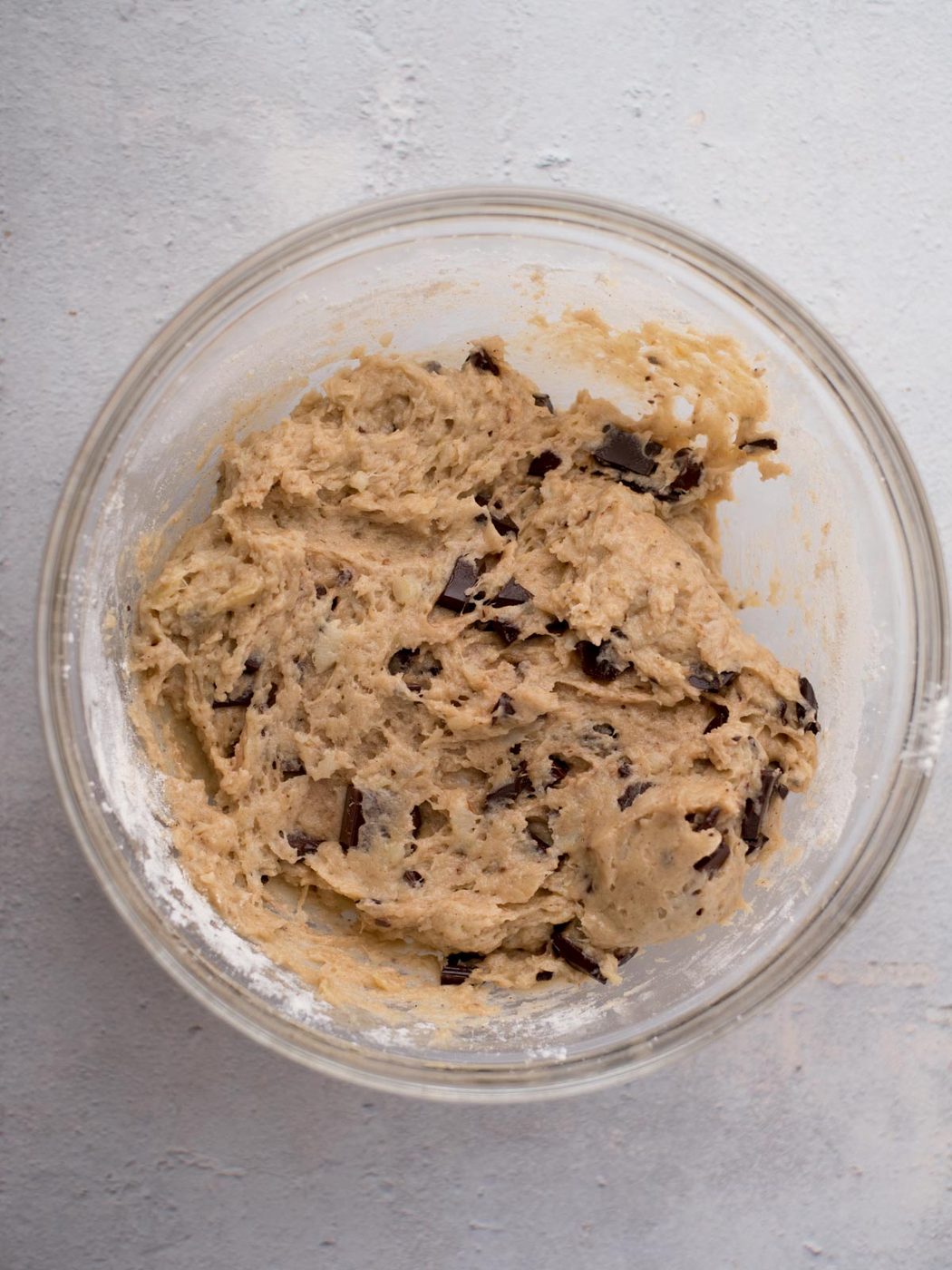 A glass bowl filled with chocolate chip cookie dough, with visible chunks of chocolate, sits on a light gray surface.