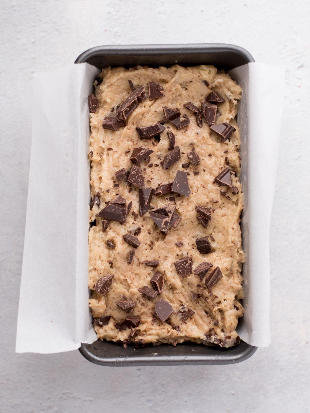 A loaf pan lined with parchment paper is filled with unbaked batter topped with chunks of dark chocolate, ready to be baked. The pan sits on a light grey surface.