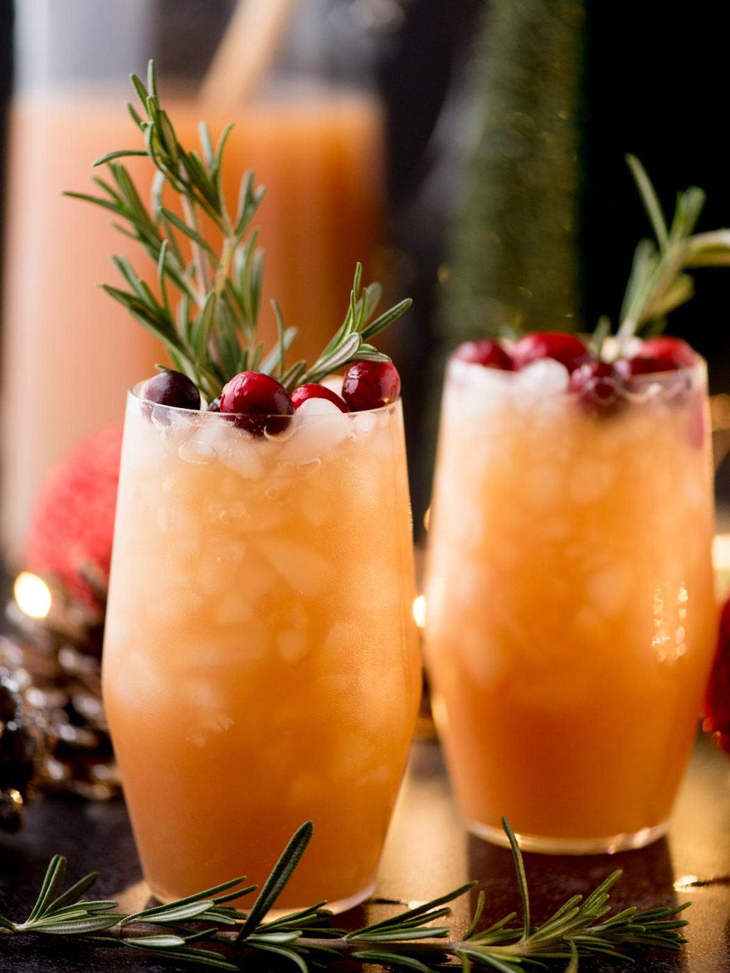 Two tall glasses filled with an orange-hued cocktail, topped with ice, cranberries, and rosemary sprigs. The background is softly blurred, featuring holiday decor with warm lighting.