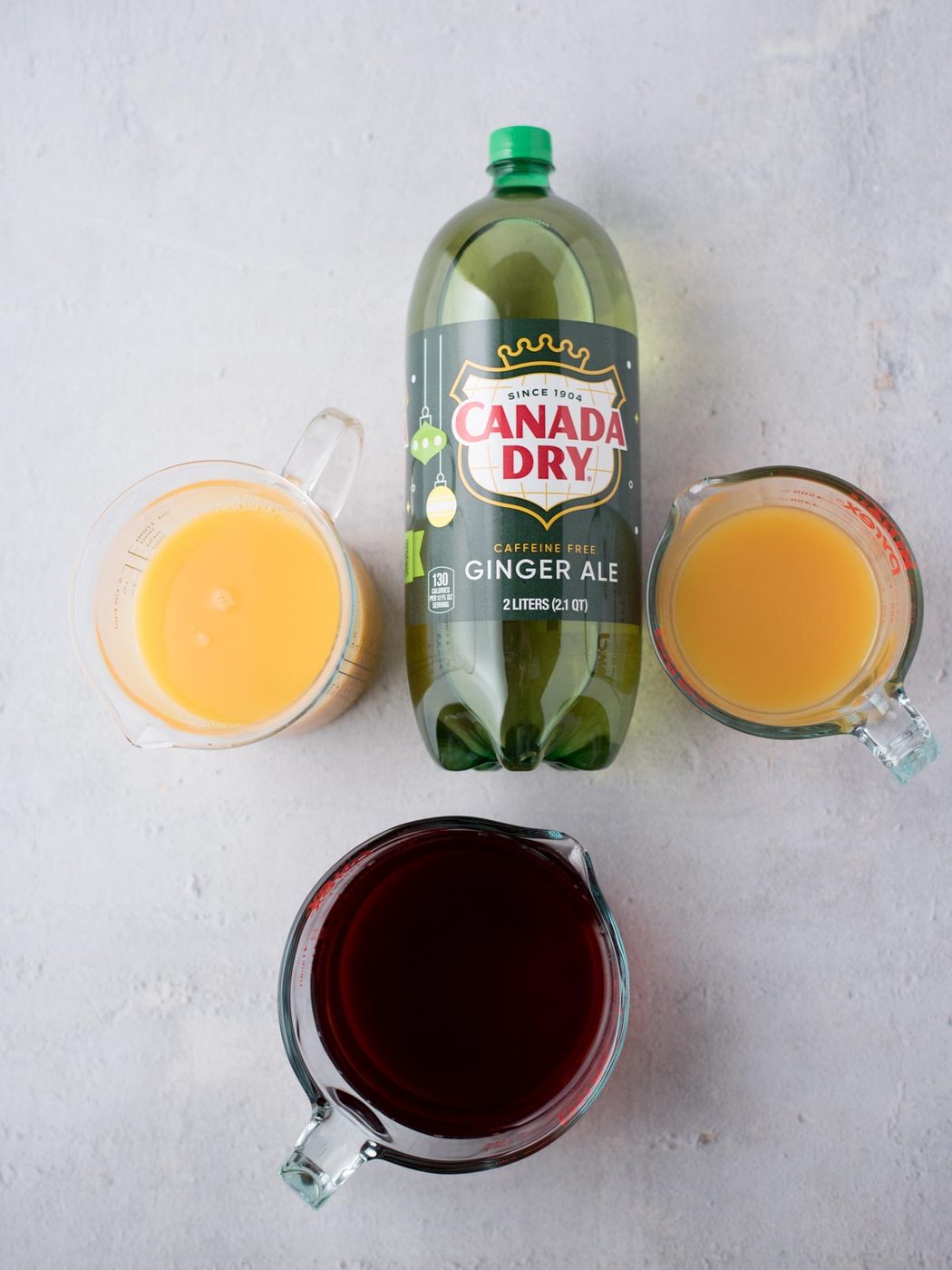 A bottle of Canada Dry ginger ale is surrounded by three measuring cups filled with different liquids: orange juice, cranberry juice, and pineapple juice. They are arranged on a light gray surface.