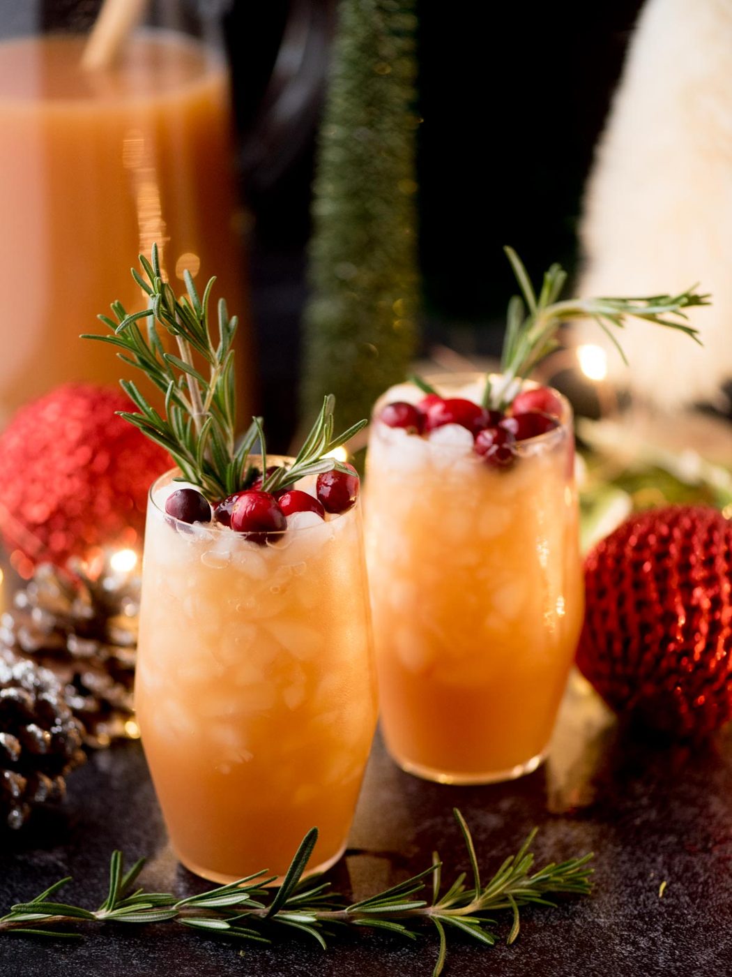 Two festive cocktails in tall glasses, garnished with rosemary sprigs and cranberries. Surrounded by holiday decor including pine cones, ornaments, and warm lights. A beverage pitcher is in the background.