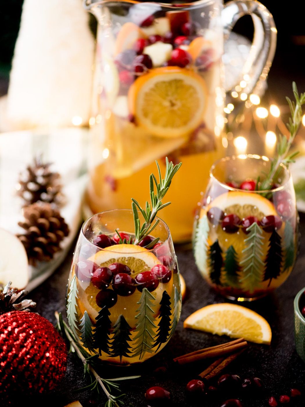 Holiday-themed sangria in pine-themed glasses, garnished with cranberries and rosemary. A pitcher with sliced fruits sits in the background, surrounded by festive decorations like pinecones, orange slices, and cinnamon sticks.