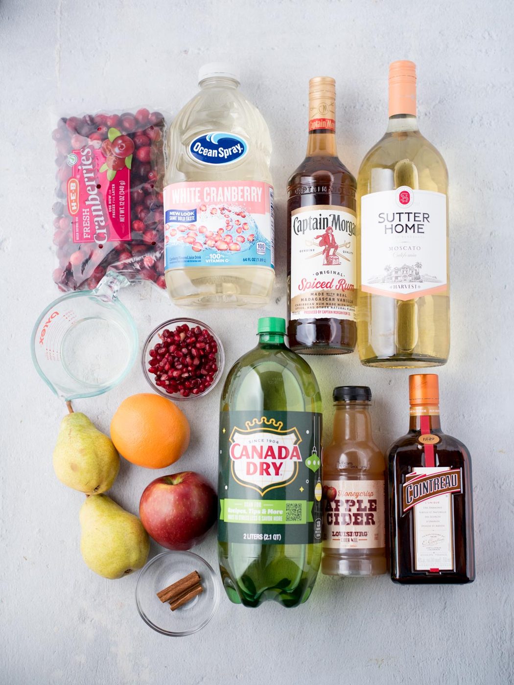 Image of various drink ingredients on a surface, including a bag of cranberries, white cranberry juice, Captain Morgan spiced rum, Sutter Home Moscato, Canada Dry ginger ale, maple cider, Cointreau, pomegranate seeds, pears, an orange, and cinnamon sticks.