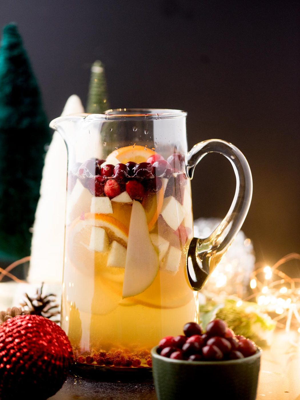 A glass pitcher filled with festive sangria, featuring slices of apples, oranges, and cranberries. It is surrounded by holiday decorations, including pine cones, ornaments, and small lit string lights.