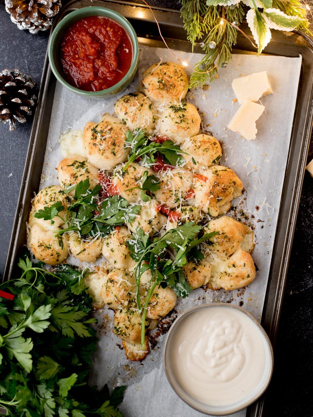 A festive, tree-shaped pull-apart bread with herbs and cheese on a baking tray, served with tomato sauce and a creamy dip, garnished with fresh parsley and Parmesan cheese. Holiday decor surrounds the tray.