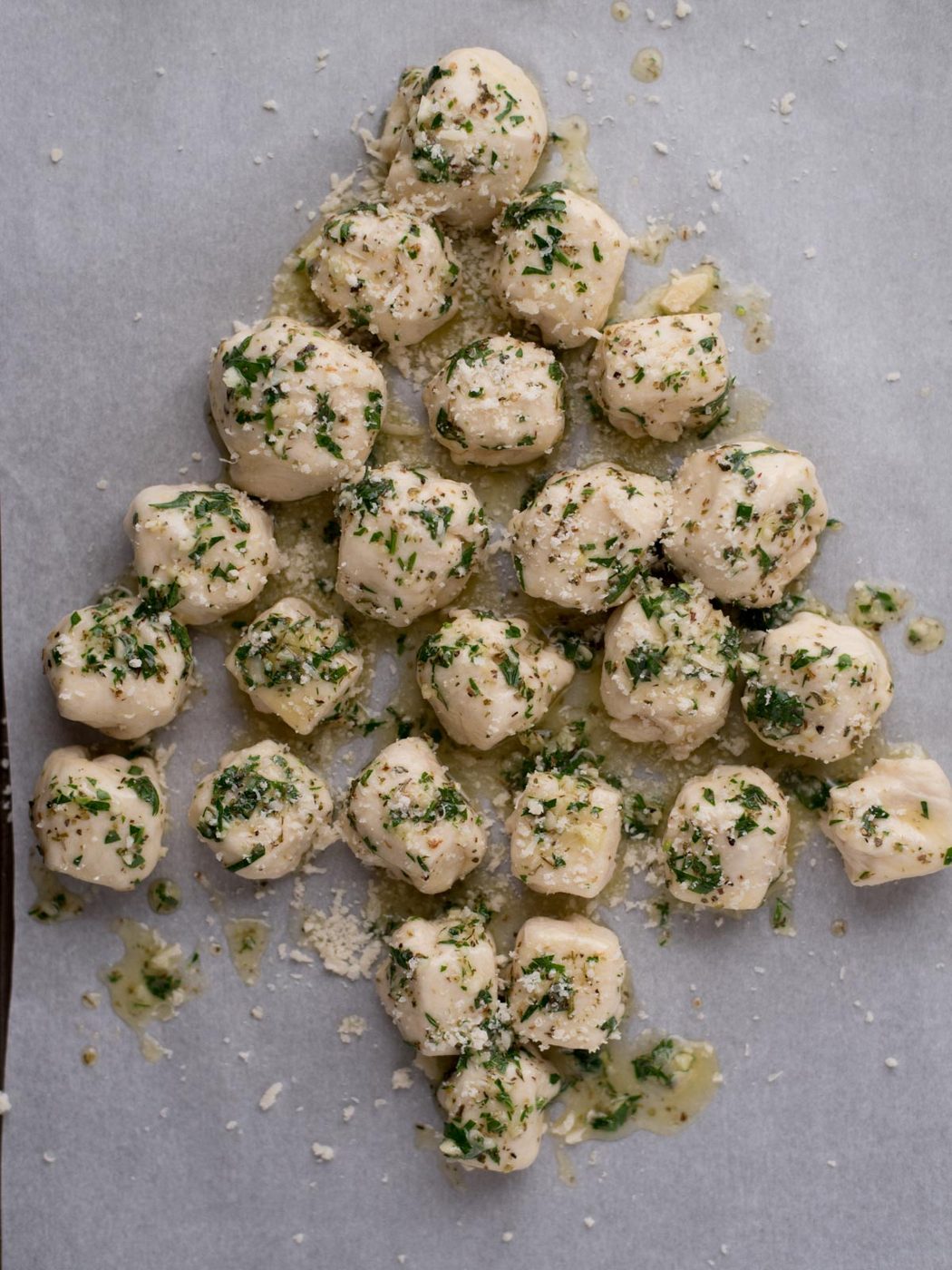 Balls of dough topped with chopped herbs and grated cheese are arranged on a baking sheet in the shape of a Christmas tree.