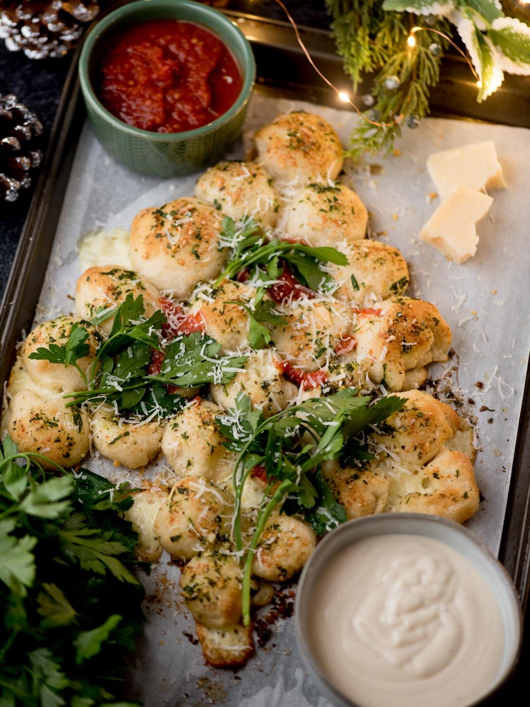 A tray of pull-apart garlic bread bites topped with herbs and grated cheese, garnished with fresh arugula, served with bowls of marinara sauce and creamy dip, and pieces of parmesan on the side.