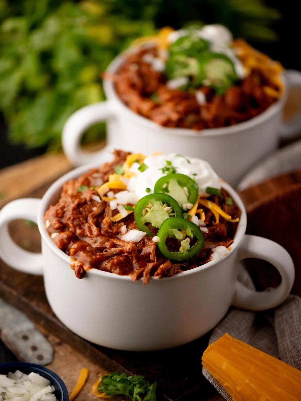 Two white bowls filled with chili, topped with sliced jalapeños, shredded cheese, and sour cream, sit on a wooden board. Fresh herbs and cheese are visible in the background.