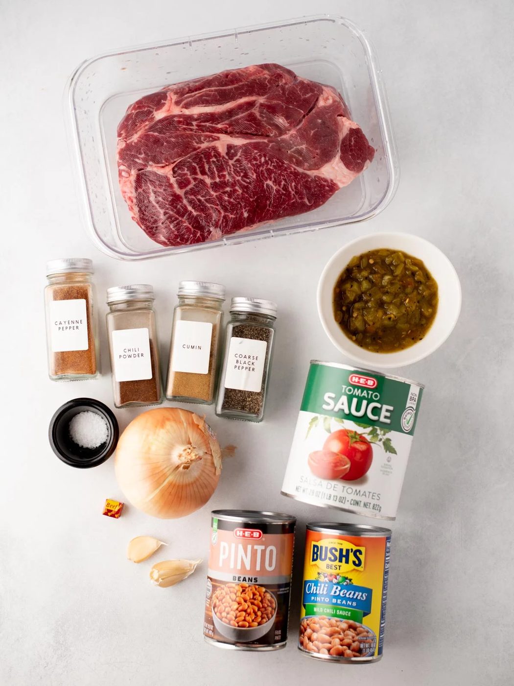 Ingredients for chili are arranged on a light surface, including a large cut of raw beef, cans of tomato sauce, pinto beans, and chili beans, spices in jars, an onion, garlic cloves, a bouillon cube, and a bowl of green chilies.