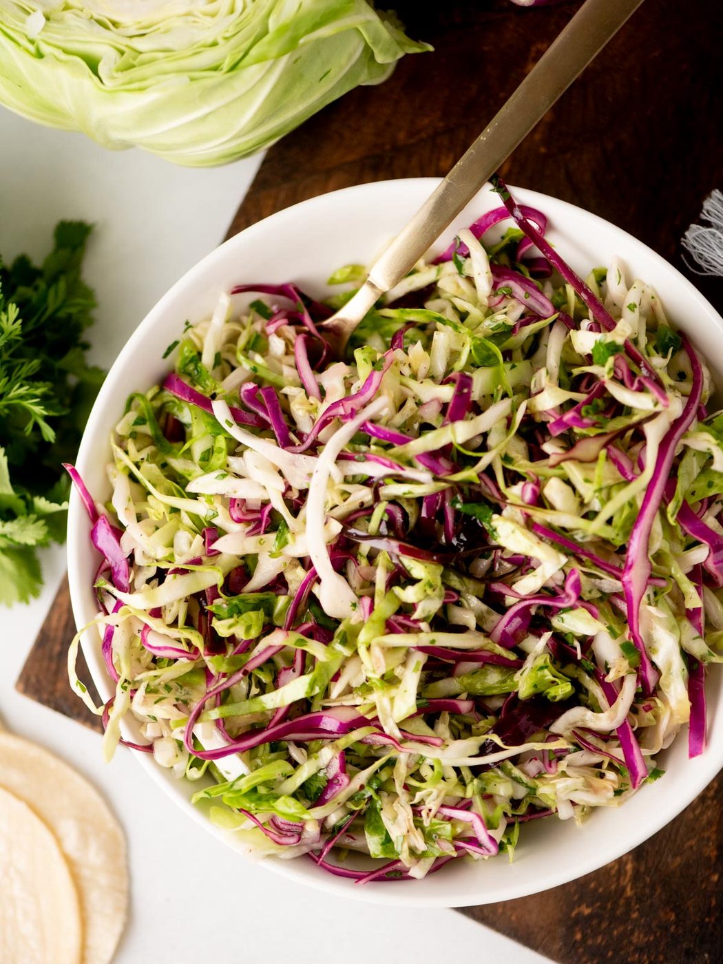 A bowl of shredded cabbage slaw with green and purple cabbage and herbs, served on a wooden surface with a gold fork. Fresh cabbage, parsley, and tortillas are visible nearby.