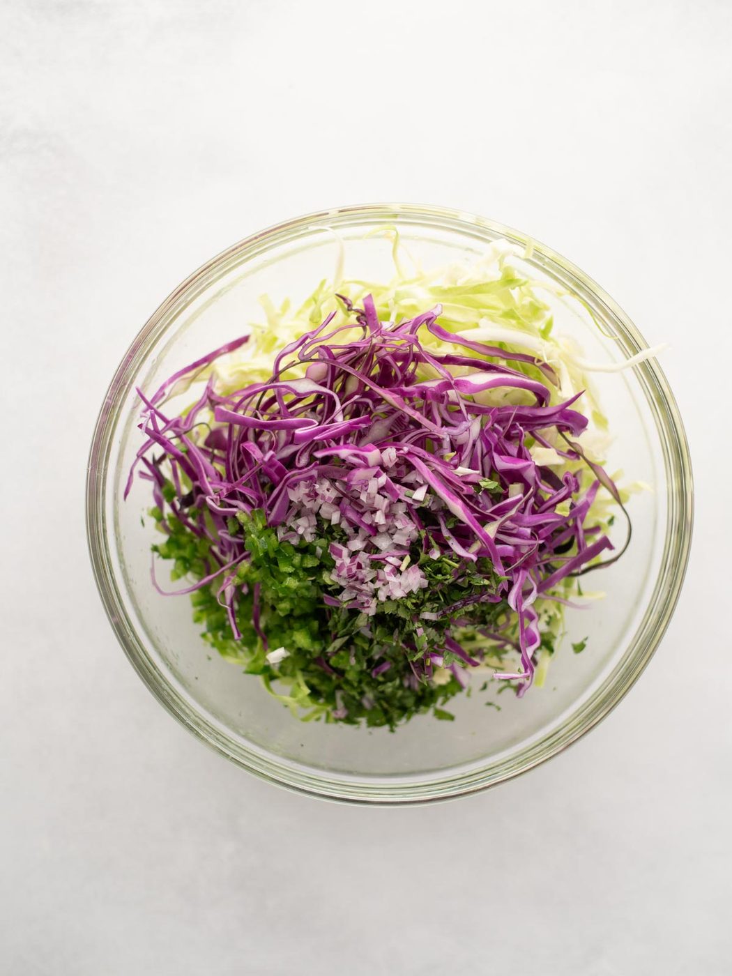 A glass bowl filled with shredded green cabbage, shredded purple cabbage, chopped cilantro, and finely diced red onion, all arranged in separate sections on a white background.