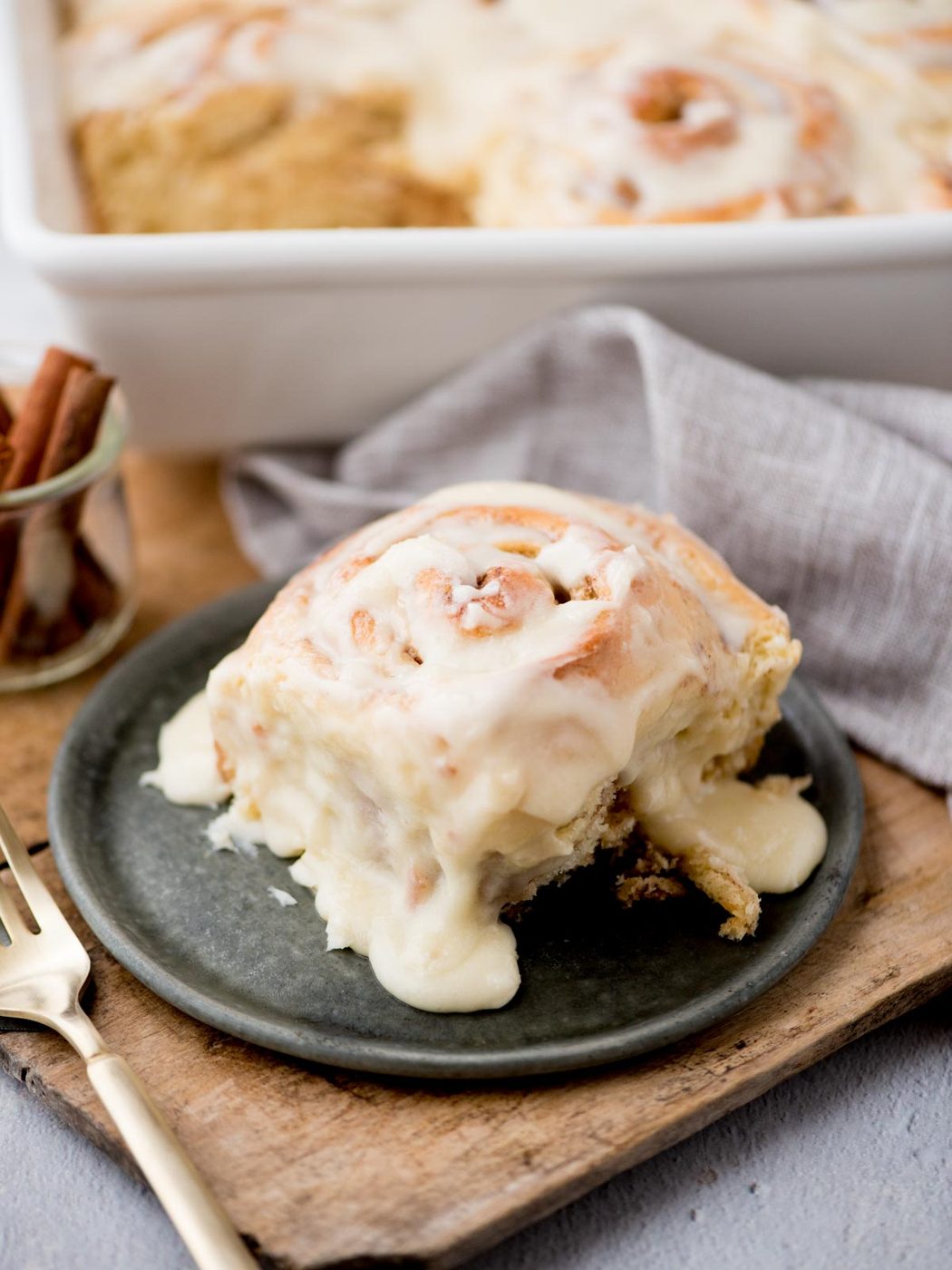 A close-up of a cinnamon roll with creamy white icing on a dark plate. A fork is placed beside it on a wooden board. In the background, theres a glass jar with cinnamon sticks and a tray of more cinnamon rolls, partly covered by a gray napkin.