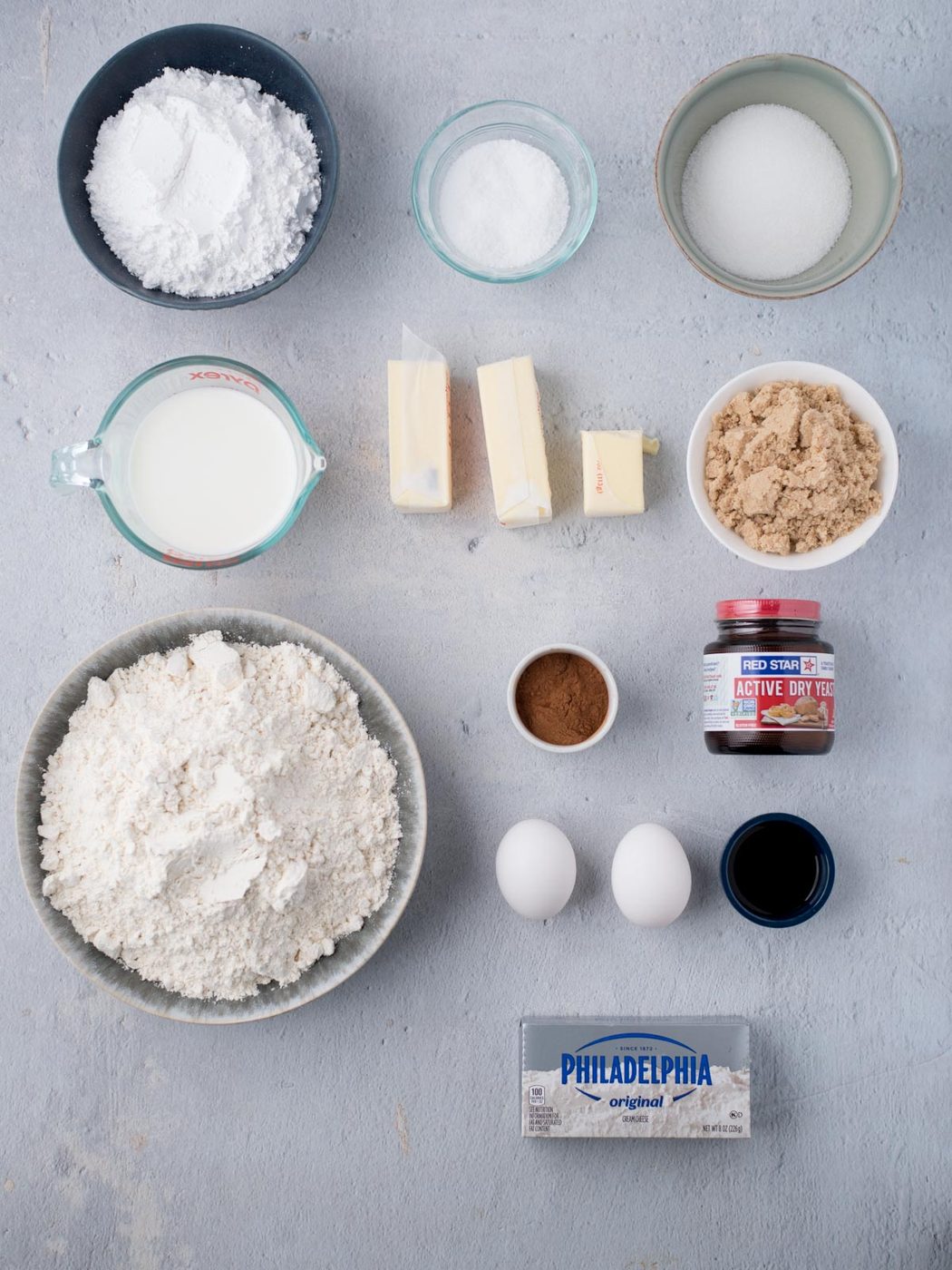 Overhead view of baking ingredients on a gray surface, including flour, eggs, butter, cream cheese, brown and white sugar, milk, active dry yeast, cinnamon, and vanilla extract, all in separate bowls or containers.