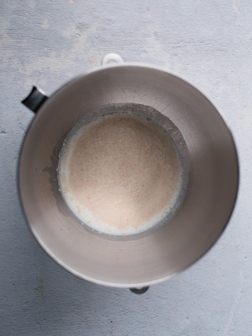A top-down view of a metal mixing bowl with frothy liquid inside, placed on a light gray surface.