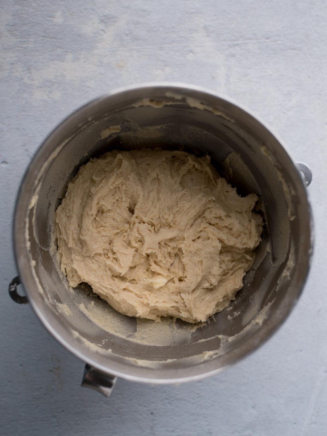 A silver mixing bowl filled with creamy, unbaked dough sits on a light gray surface. The dough has a slightly rough texture, with streaks of flour visible on the sides of the bowl.