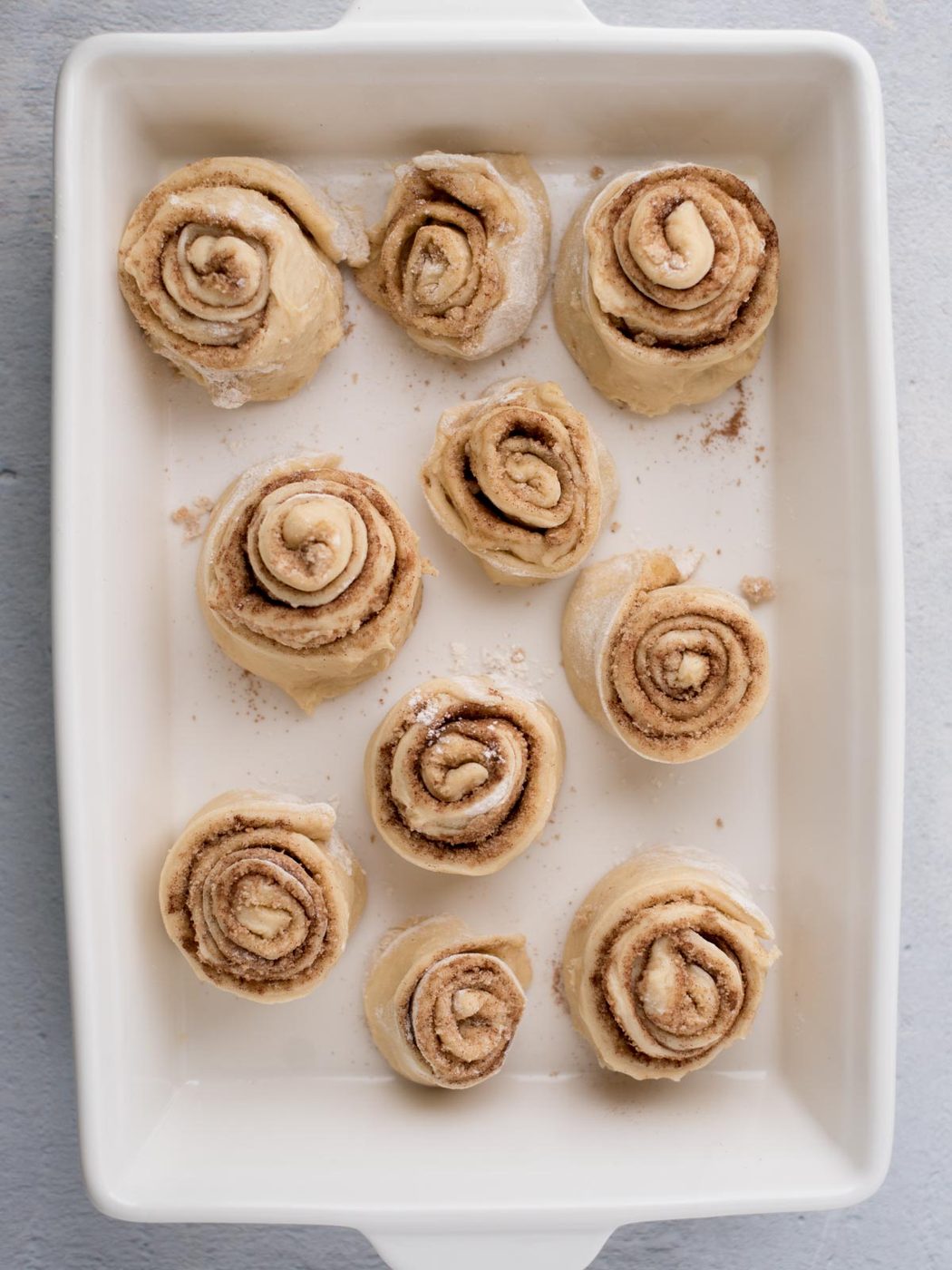A baking dish with ten uncooked cinnamon rolls spaced evenly apart. The rolls are spiraled with visible layers of dough and cinnamon-sugar filling, ready to be baked.