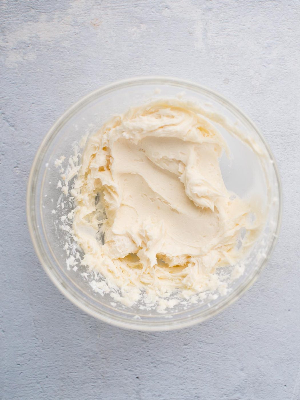 A clear glass bowl filled with creamy, smooth white frosting on a light gray textured surface. The frosting appears to have a soft, whipped consistency with gentle peaks and swirls.