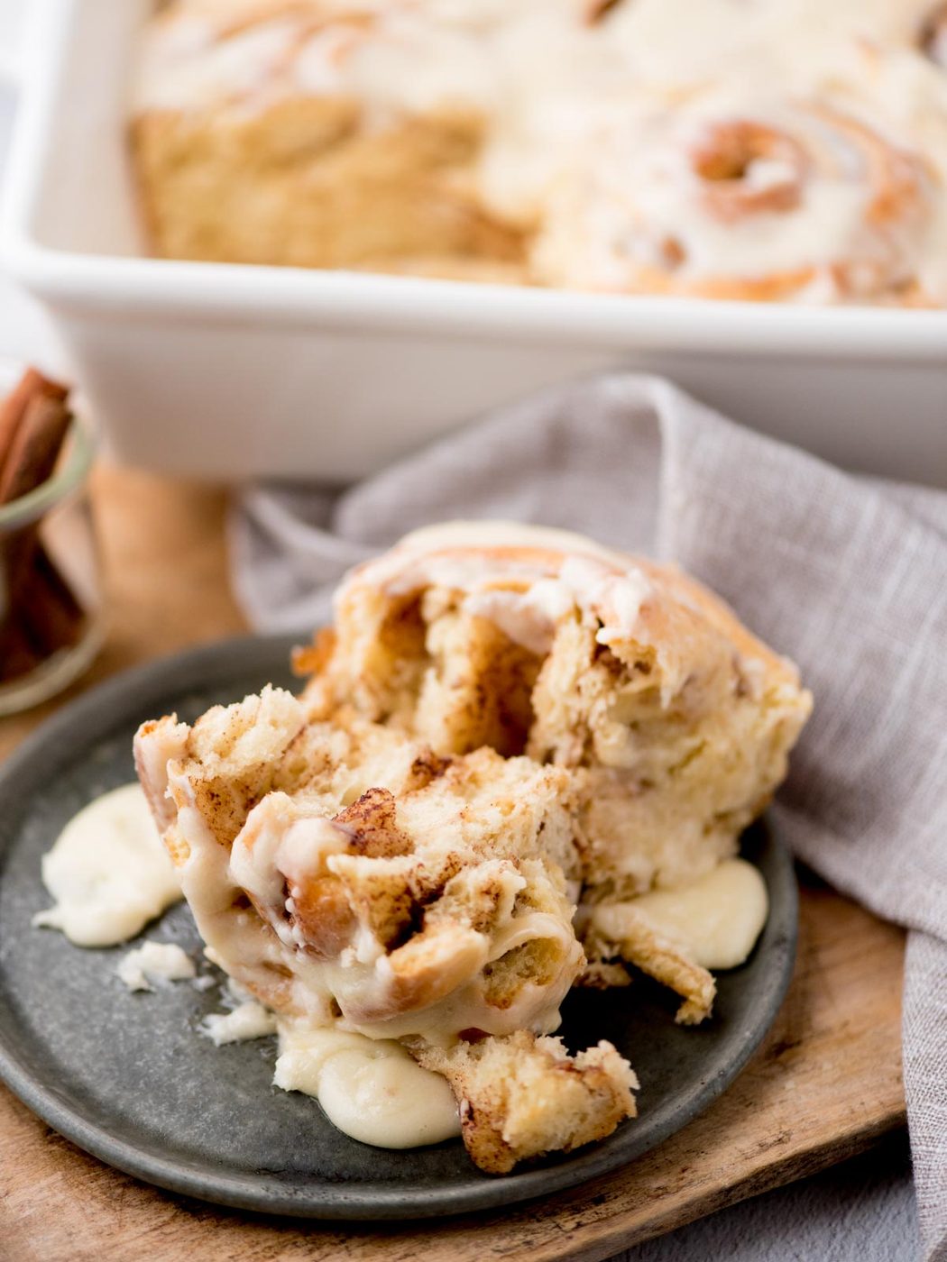 A freshly baked cinnamon roll is on a plate, topped with creamy icing. Its partially pulled apart, revealing a soft, fluffy interior. In the background is a baking dish with more cinnamon rolls and a jar of cinnamon sticks. A cloth napkin is draped nearby.